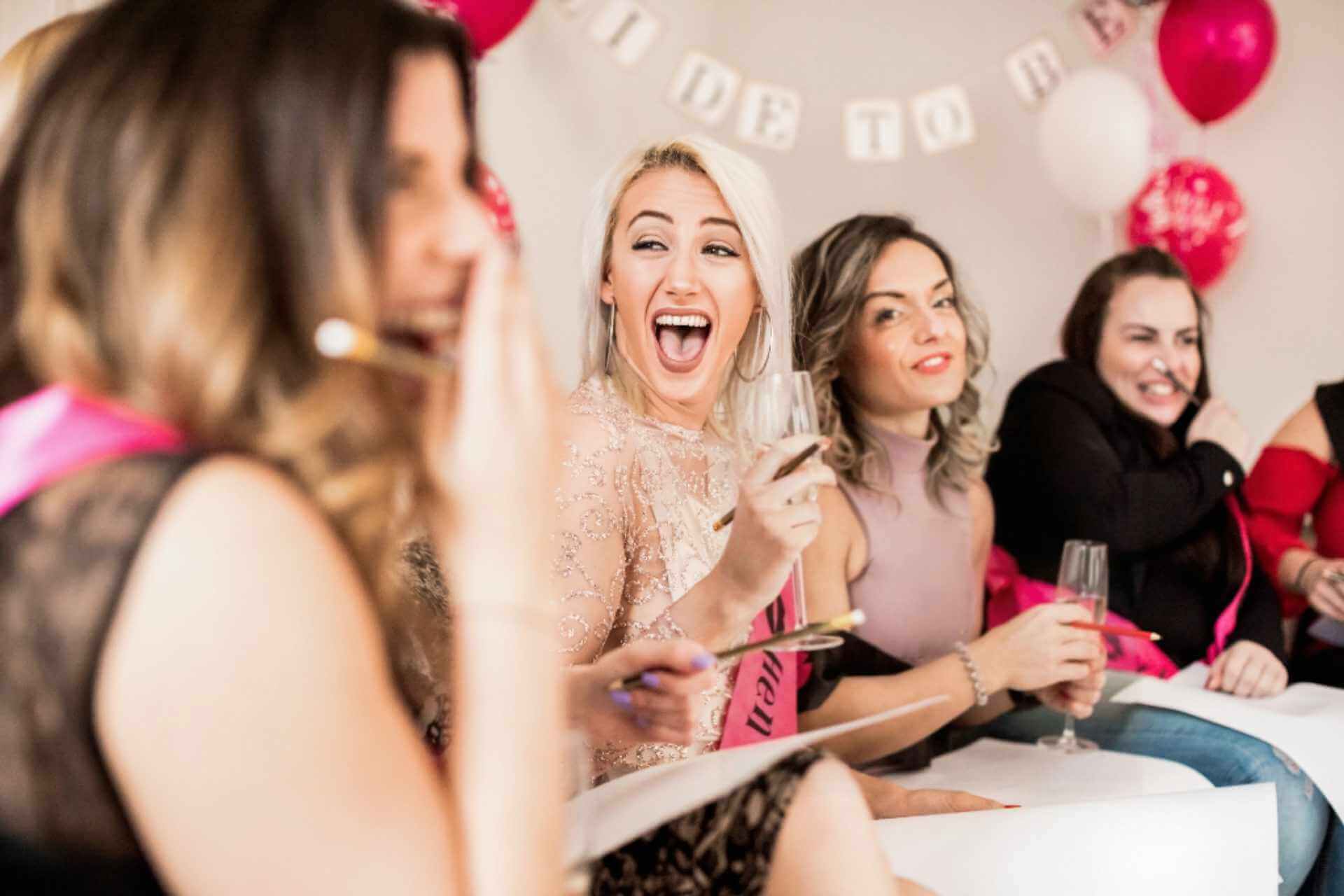 Excited hen party guests laughing and sketching during a fun life drawing session, with pink sashes, balloons and prosecco – the perfect cheeky activity with Butlers with Bums.